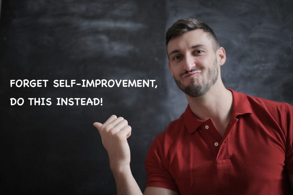forget self-improvement, do this instead! A man in red shirt pointing his right thumb at the chalkboard behind him.